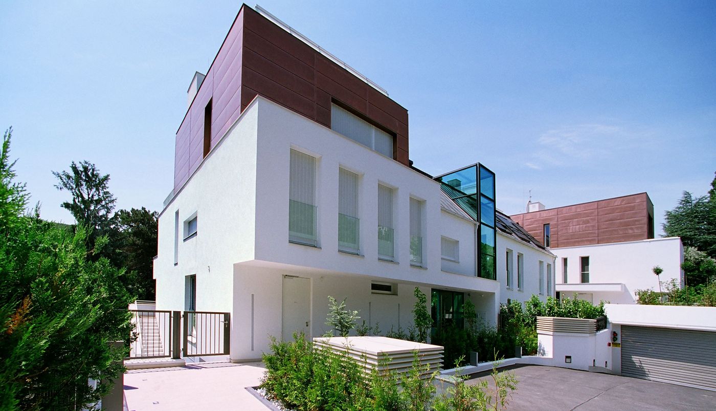 Foto: Wohnhausanlage mit zweigeschossigen Gebäuden, hohen Fenstern und verglastem Stiegenhaus; im Vordergrund die Einfahrt in eine Tiefgarage und Grünflächen