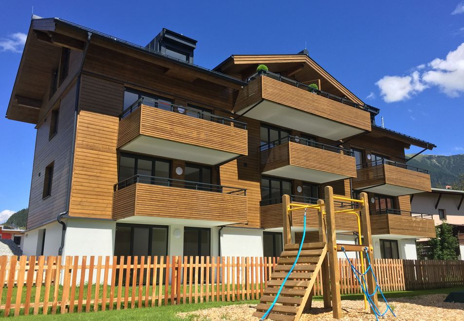 Visualisierung: Wohnhaus mit Balkonen und Holzfassade in hellem Sonnenlicht; im Vordergrund ein Spielgerät für Kinder auf grüner Wiese