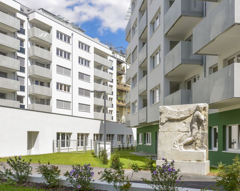 Photo: bas-relief of a man sculpted into a block of stone placed on a plinth; in the foreground, a green, fenced-in lawn bordered by ornamental shrubs; in the background, the façades of two deeply aligned residential buildings